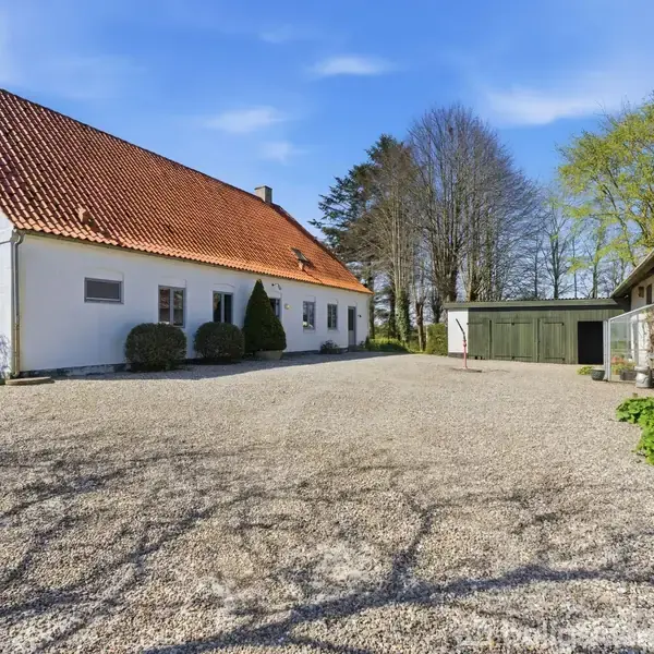 Hvid bygning med rødt tegltag står på en grusbelagt gårdsplads, omgivet af grønne træer og en mindre bygning. Solrigt vejr, med klar blå himmel.