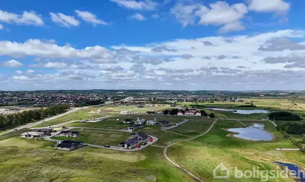Huse ligger på en åben eng med stier, omgivet af marker og en lille sø. Byen ses i horisonten under en blå himmel med skyer.