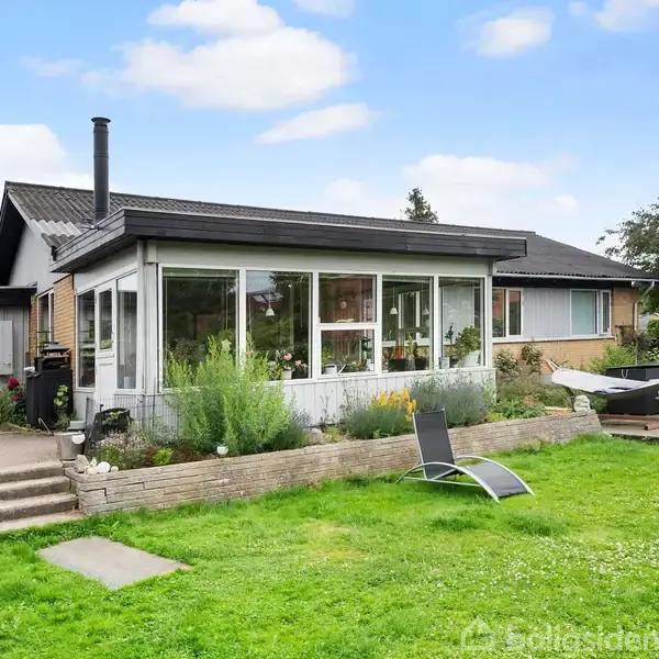 En enfamiliebolig har en vinterhave med store vinduer. Der er havemøbler og en liggestol i den grønne have. Huset står i et villakvarter under en let skyet blå himmel.