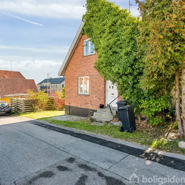 Rødt murstenshus dækket af grønne planter ved siden af en rolig vej med parkede biler og et træ. Baggrunden har flere huse under en klar blå himmel.