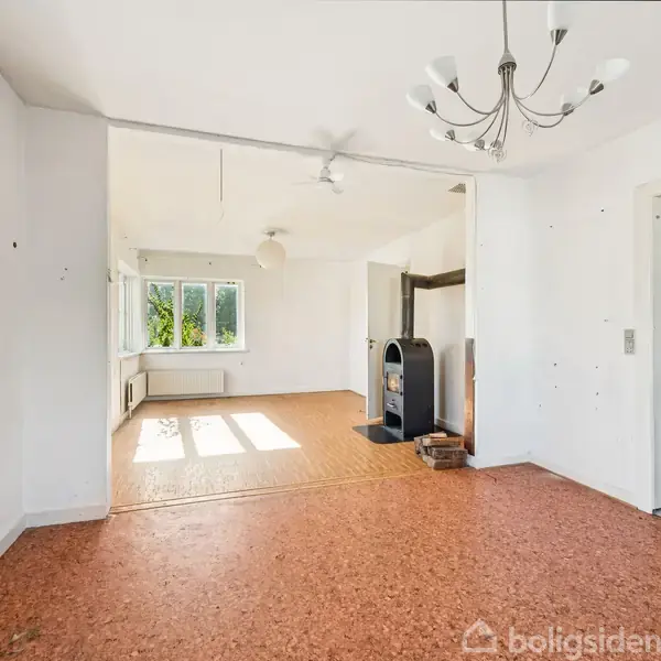 Stue med brændeovn står i et lyst rum med lyse vægge. En lysekrone og en åben dør skaber en rummelig atmosfære. Vinduer tillader naturligt lys at strømme ind.