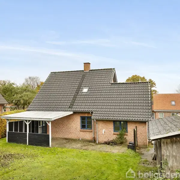 Murstenshus med mørkt tag, står i en grøn have. Der er en udestue på siden og en træbygning til højre. Himlen er blå med spredte skyer.