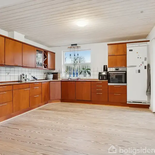 Køkken med træskabe og skuffer langs væggene. Rummeligt og lyst med hvidt loft og trægulv. I baggrunden ses et vindue, en opvaskemaskine og hvidevarer, inklusive et køleskab.