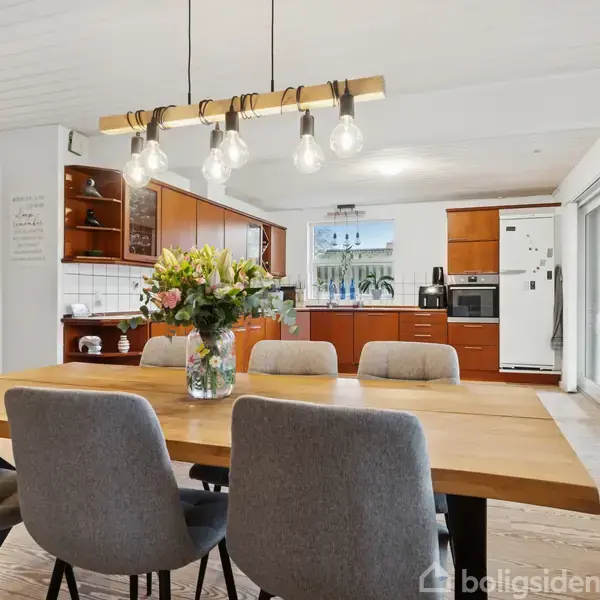 Et spisebord med grå stole står i et køkken med hvide vægge og træmøbler. En buket blomster i en vase står på bordet. Loftlampe med pærer hænger over bordet.