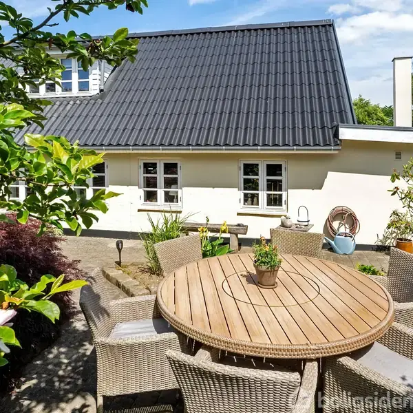 Rundt havebord i flettet materiale er omgivet af stole og har en lille plante på toppen. Det står på en terrasse foran et hvidt hus med sort tag og grønne omgivelser.