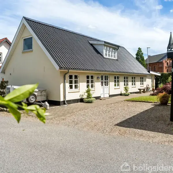 Et hvidt hus med sorte tagsten står på en grusbelagt grund. En grusvej løber foran huset, omgivet af grønne planter og flere bygninger, inklusive en kirke i baggrunden.
