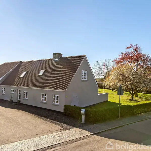 Et gråt, enfamiliehus med flere vinduer og en mørk tagbeklædning står langs en vej. Området er solrigt med en græsplæne og blomstrende træer.