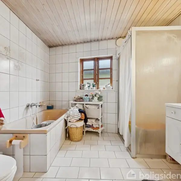 Et badeværelse med hvidt flisegulv og træloft. Et badekar med en rød nisse, en brusekabine, og en hvid vask ved siden af et forrådsskab. En kurv står ved et lille vindue.