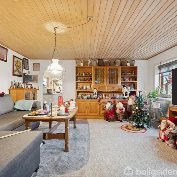 En stue med en grå sofa og et træbord, omgivet af julepynt som nisser og et lille juletræ. Træloft og skab fyldt med dekorative genstande fuldender scenen.