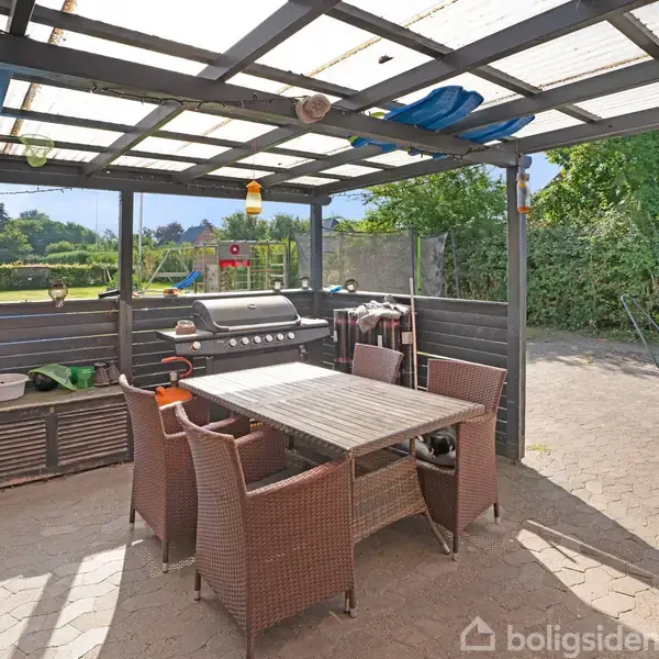 Grill står under et overdækket terras, omgivet af rattan-stole og et bord i en have med grøn bevoksning. Sollys skinner ind gennem det gennemsigtige tag.