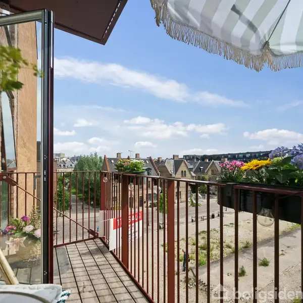 Balkon med farverige blomster arrangeret langs gelænderet under en stribet parasol. Solskinsdag med udsigt over byens tage og en åben grusplads i baggrunden.