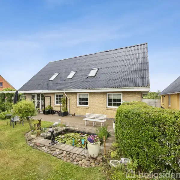 Hus med sort, skråt tag og beige murstensvægge står i en velplejet have med flisebelagt terrasse. Omkring ses græsplæne, buske og en bænk. Skyfri blåt himmel i baggrunden.