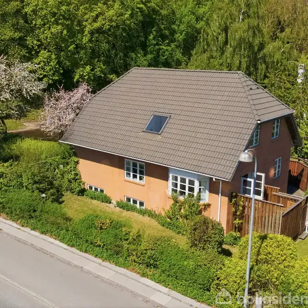 Et rødt hus med en gråt tag står omgivet af frodig have og blomstrende træer. En vedligeholdt vej og en lygtepæl er i forgrunden, hvilket skaber en fredelig, forstadslignende atmosfære.