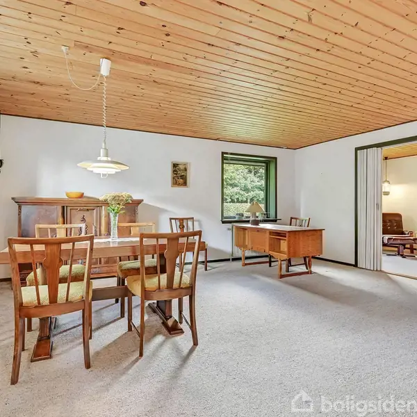 Spisebord med seks stole står i en spisestue med træloft og tæpper. Bordet er dækket med en vase blomster. Til højre ses en åben stue med lænestole.