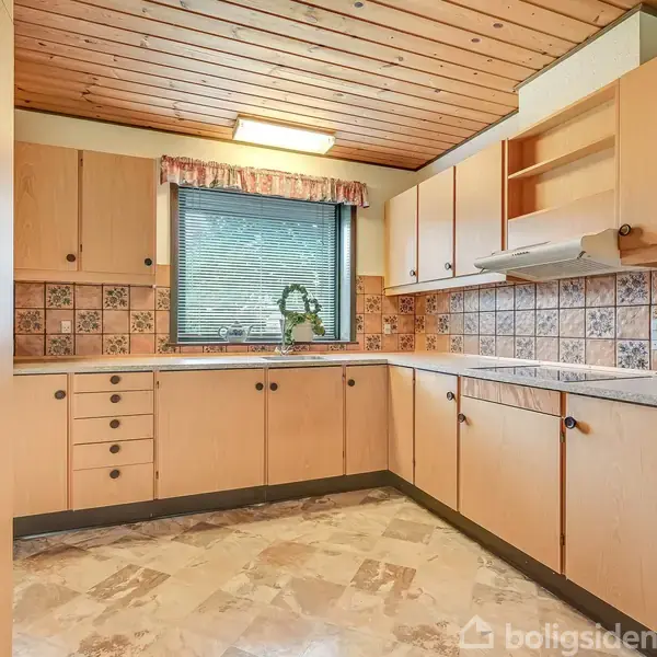 Køkken med træskabe og modernistisk flisemønster, stående urørt. Et vindue med persienner lader dagslys komme ind, mens lysebrune møbler og marmorlignende gulv definerer rummet.