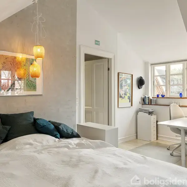 En stue indeholder en grå sofa med farverige puder, retter sig mod et sofabord med en blomstervase. En lænestol er placeret ved siden af. Væggene har kunstværker, og et vindue tilføjer naturligt lys. En fladskærms-TV står til højre.