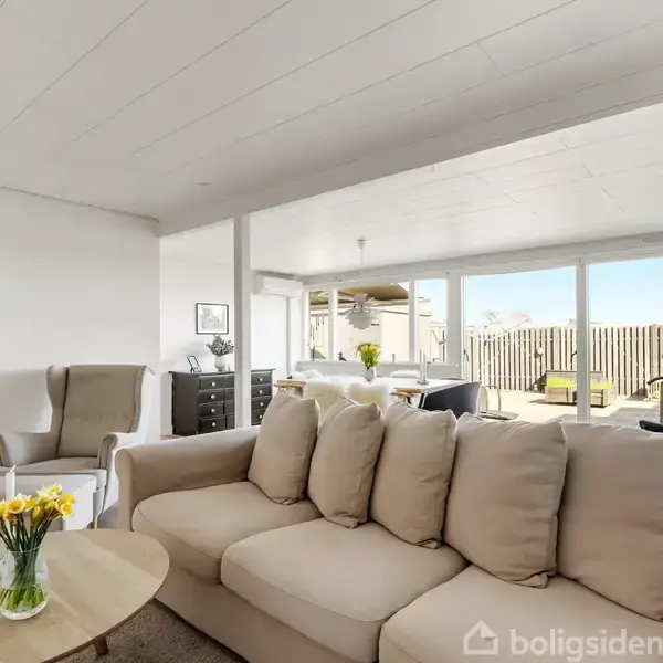 En lys stue med en beige sofa står foran store vinduer. Et rundt sofabord med blomster er midtpunktet. Til venstre er en lænestol og et vitrineskab.