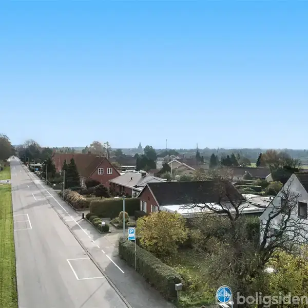 En stille villavej strækker sig langt, flankeret af velplejede huse og haver. Træer og buske omkranser området, mens en blå cykelsti-skilt står tydeligt langs fortovet.