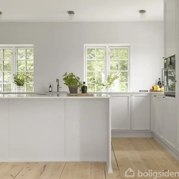 Et lyst køkken med en hvid køkkenø, hvor der står planter og flasker. To vinduer i baggrunden giver udsigt til grønne træer, og gulvet er lyst træ.