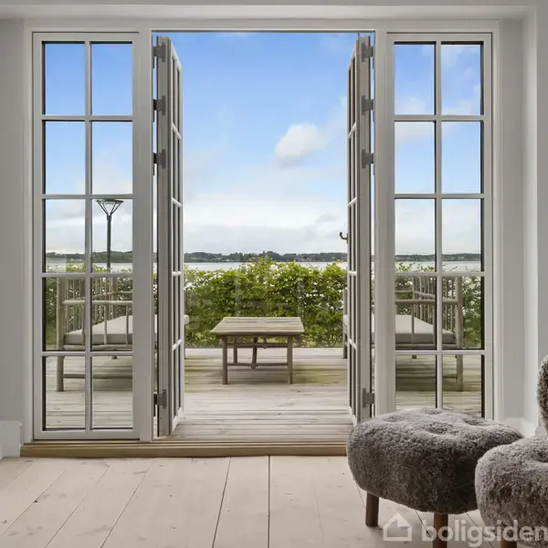 Dobbelt fransk dør åbner ud til en terrasse med stole og et bord, omgivet af grøn beplantning og udsigt til en klar blå himmel. Indendørs er der komfortable stole.