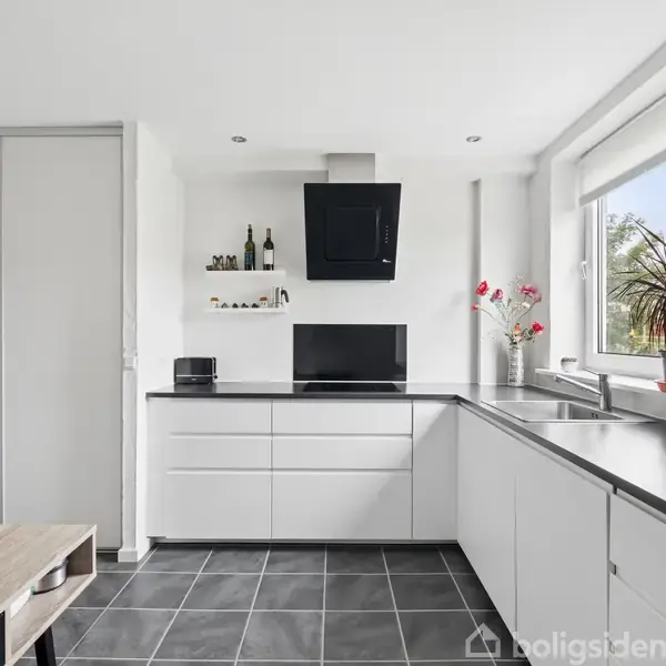 Køkken med sorte fliser, hvide skabe og sort emhætte. Bordet står modsat en stor vindueskarm med planter og blomster. Lysindfald fra højre giver et lyst og moderne udtryk.