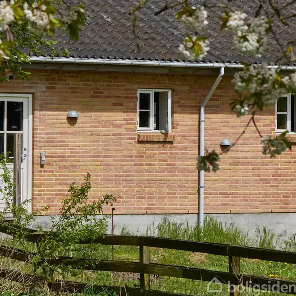 En murstensbygning med en dør og to vinduer. En træhegn og grønne planter omgiver huset. Blomstrende grene hænger ned foran bygningen.
