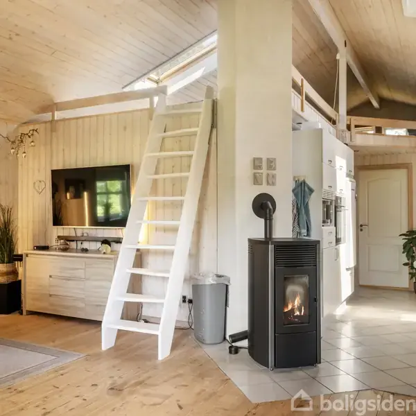 Stue med en hvid trappe fører til hems, en brændeovn ved siden af til venstre, sofa og TV-bord. Lys trævægge og lofter skaber en hyggelig atmosfære.