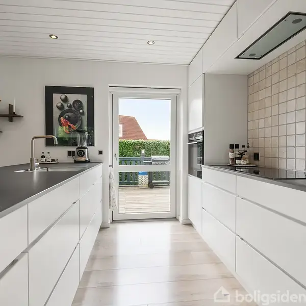 Et moderne køkken med hvide skabe strækker sig langs to vægge. En glasdør fører ud til en terrasse med udsigt til en rød bygning. Spisebord og kunst på væggen.
