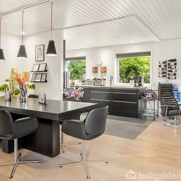 Sort spisebord med seks stole står i en lys spisestue. Tre lamper hænger over bordet. I baggrunden ses et moderne køkken med vinduer og farverig kunst på væggene.