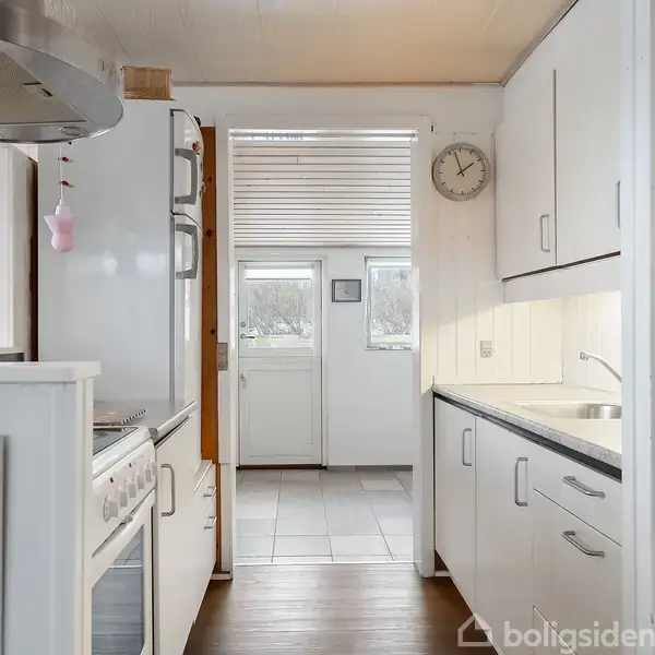 Et hvidt, smalt køkken fører ind til et rum med flisegulv. Et ur hænger på væggen, mens køkkenredskaber og grønne flasker er synlige på hylderne.