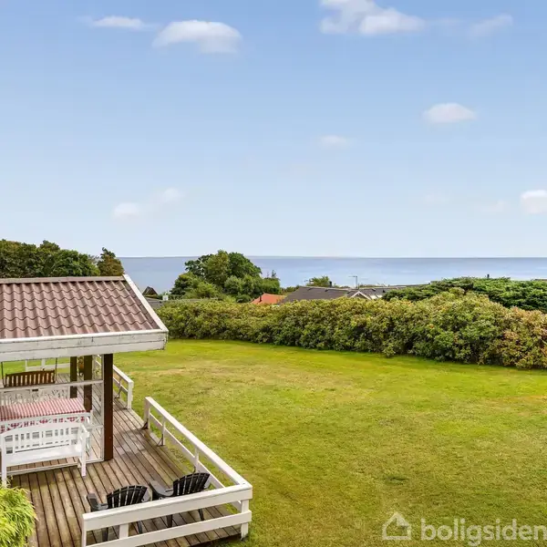 Et træhus står stille ved en frodig grøn have med udsigt over havet. Omkring huset ses buske og træer under en klar blå himmel.