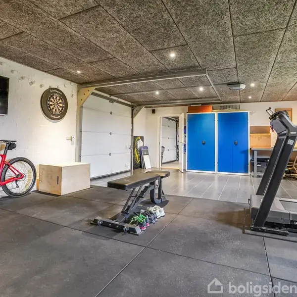 Et træningslokale med en rød cykel, en bænk, og et løbebånd i aktion. Omgivelserne inkluderer en dartskive, en tv-skærm og skabe. Træningsudstyr er spredt rundt i rummet.