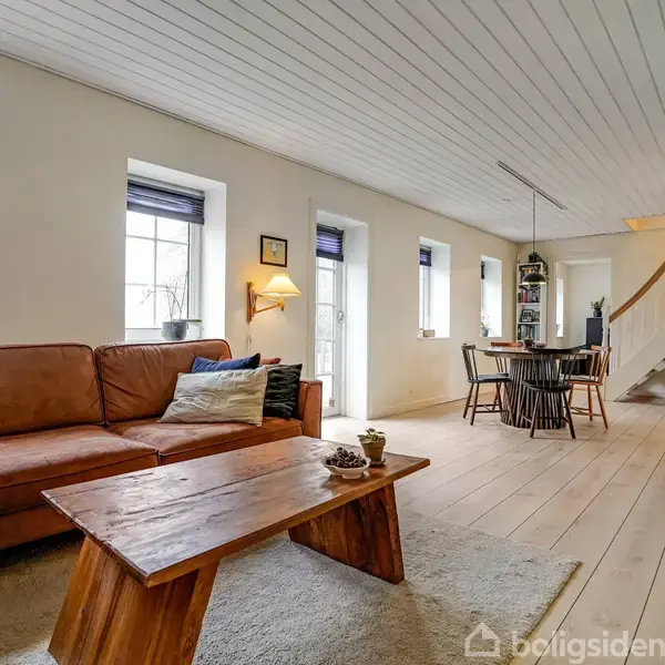 Brunt sofaarrangement står i en lys dagligstue med hvidmalet loft, træbord og tæppe. Spisebord og stole er placeret nær trappe. Flere vinduer giver naturligt lys.