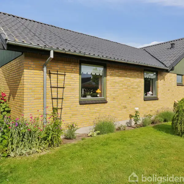 Et gult murstenshus med sort tag står i en velholdt have med grøn plæne og blomsterbede. Der er flere vinduer langs husets side, og det er en solrig dag.