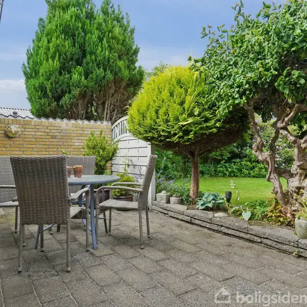 Terrassemøbler står på en flisebelagt terrasse. Ved siden af er der velplejede buske og et lille træ. En murstensmur omkranser området, mens en græsplæne er synlig i baggrunden.