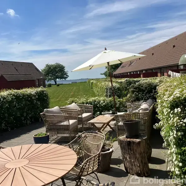 Terrassemøbler står på flisebelagt terrasse. Parasol skygger over borde og stole. I baggrunden er der grønne buske, græsplæne og huse med skrå tage under en blå himmel med skyer.