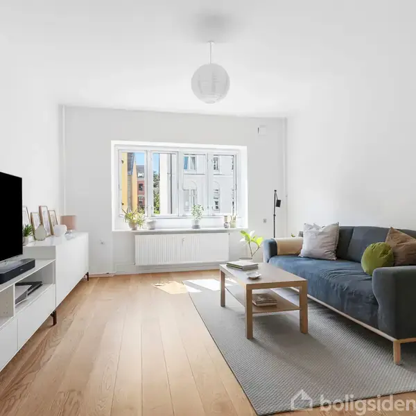 En grå sofa står på et tæppe, omgivet af planter og møbler i en lys stue med trægulv. Store vinduer lader dagslys strømme ind, hvilket skaber en indbydende atmosfære.