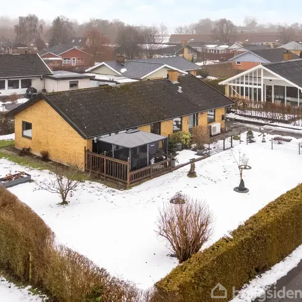 Et gult murstenshus står i en sneklædt have med hæk omkring. Der er en træterrasse foran huset og sne dækker gangarealerne, mens nabohuse ses i baggrunden.