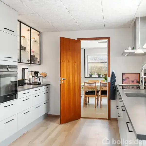 Køkken med hvide skabe og sorte håndtag har integrerede apparater og kaffemaskine på bordpladen. Dør i træfarve åbner til spisestue med træbord og stole.