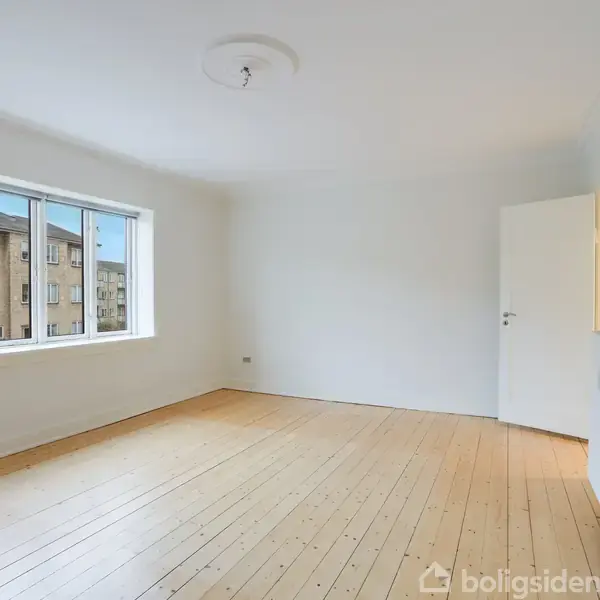 Et lyst, tomt værelse med trægulv og hvide vægge. Et stort vindue lader naturligt lys strømme ind, og en radiator er monteret nær en åben dør til højre.