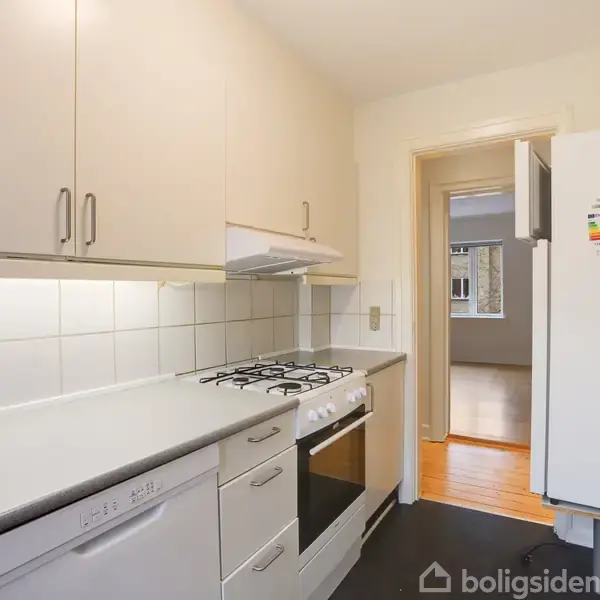 En lys køkken indretning, med gaskomfur og ovn i midten, står op mod lyse skabe. Det fører ind til en stue med trægulv, belyst udenfra.