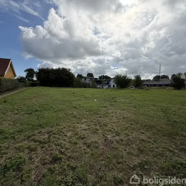 En græsmark strækker sig foran flere boliger. Markens korte græs viser, at den er nyligt slået. Huse og træer omkranser området under en dramatisk skyfyldt himmel.