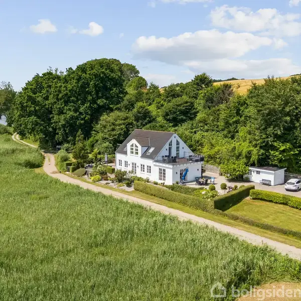 Et hvidt hus med sort tag står ved siden af en sø. Det er omgivet af grønne træer og græs. En grusvej løber langs ejendommen.