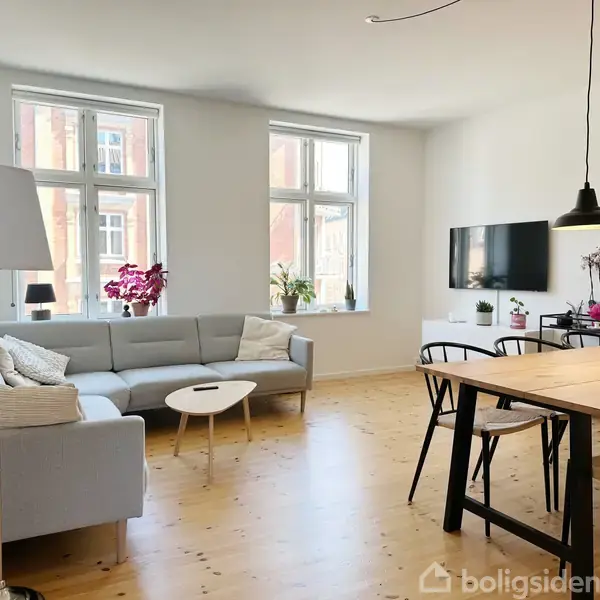 Lys stue med grå hjørnesofa, stuebord, og lamper. Spisebord med sorte stole til venstre; fladskærm hænger på væggen. Store vinduer giver udsigt og naturligt lys.