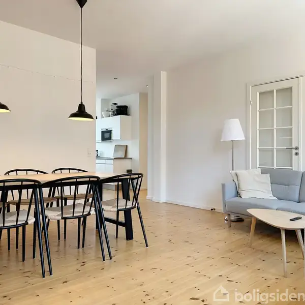 Spisebord med seks stole står i en stue med trægulv. To hængelamper lyser over bordet. I baggrunden ses en grå sofa ved siden af dobbeltdøre.