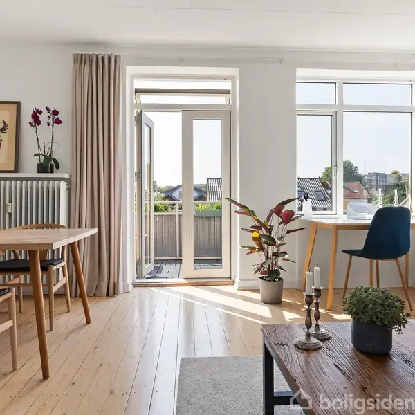 Stue med lyse trægulve og spisebord, sofa og sofabord. En dør fører til en balkon. Store vinduer giver udsigt til bygninger. Plante og stearinlys pynter rummet.