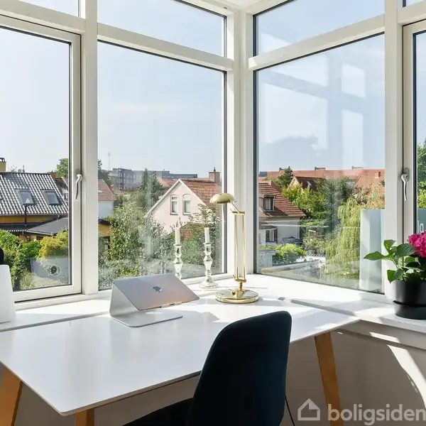 Bord ved stort vindue med laptop, lampe og plante. Udsigt til nabolag med huse og haver under blå himmel.