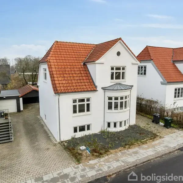 Hvidt hus med rødt tag står på flisebelagt indkørsel, omgivet af lignende boliger. Foran ses en vej og en mindre have med blomsterbede.