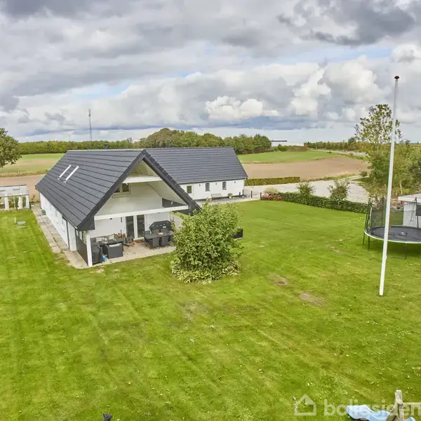 Et moderne hus med sort tag ligger på en velplejet græsplæne. Til højre er der en trampolin og en flagstang. Omgivet af åbne marker og træer under en overskyet himmel.