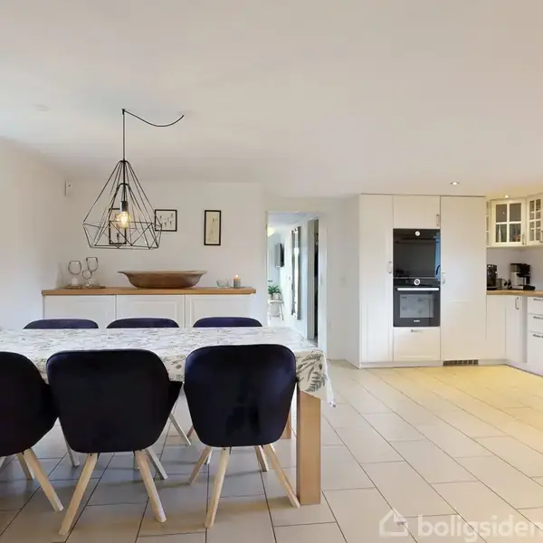 Et spisebord med mørke stole står i køkkenet. Bordet er dækket med dug, og over det hænger en dekorativ lampe. Køkkenet er lyst og rummeligt med hvide skabe.